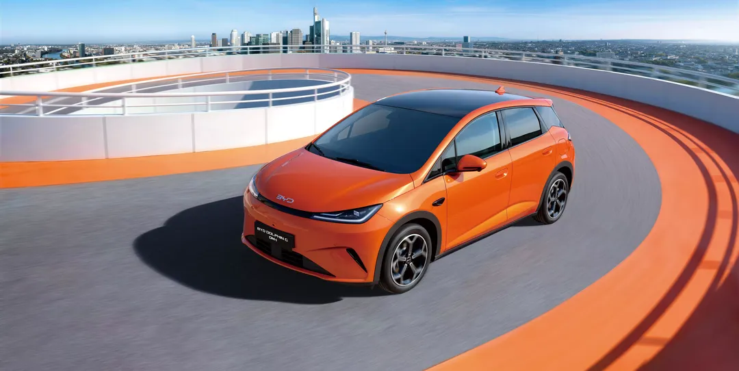 BYD Dolphin G DM-i — przód 3/4 z góry, instalacja promocyjna z pomarańczowym torem i panoramą miasta