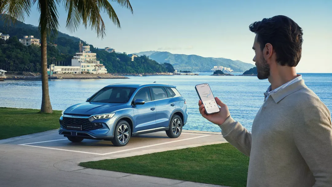 BYD SEALION 5 DM-i — przód 3/4 w kolorze niebieskim, lifestyle z mężczyzną trzymającym smartfon, tło nadmorskie