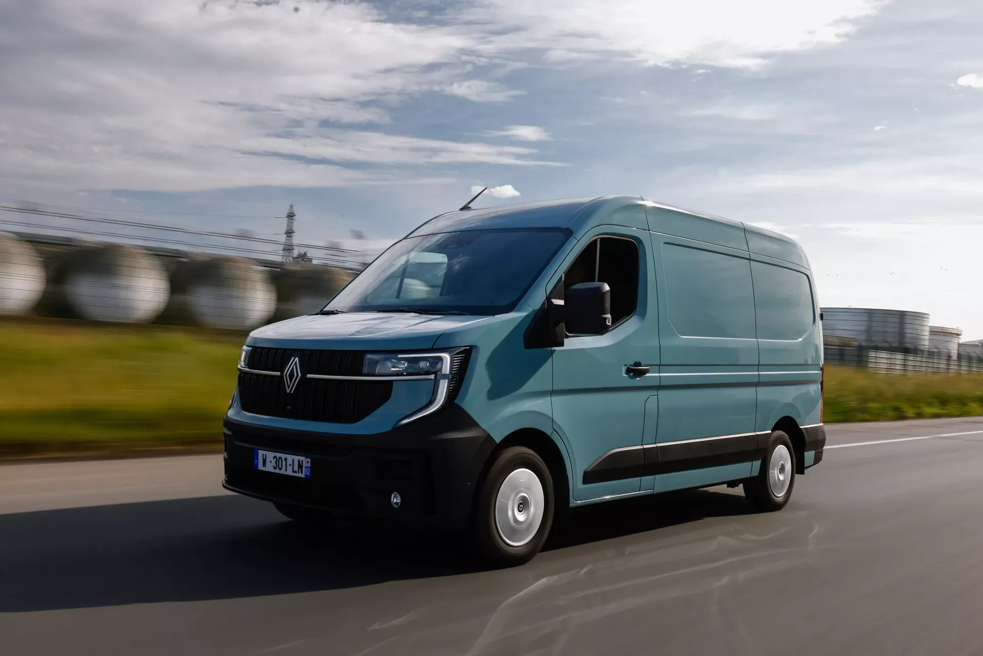 Renault Master (furgon, kolor błękitny) — przód, jazda drogą przy zbiornikach przemysłowych