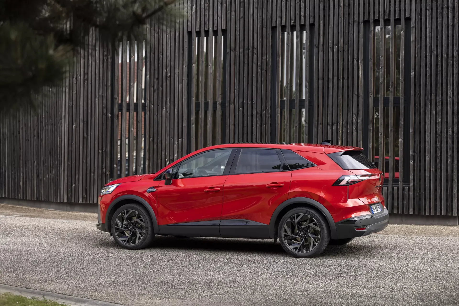 Renault Symbioz — bok z widocznym tyłem, czerwony SUV na tle drewnianej elewacji