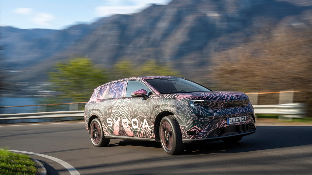 Škoda Peaq — prototyp w kolorowym kamuflażu ze wzorem fal, dynamiczne ujęcie 3/4 z przodu-lewej na alpejskiej drodze z górami w tle